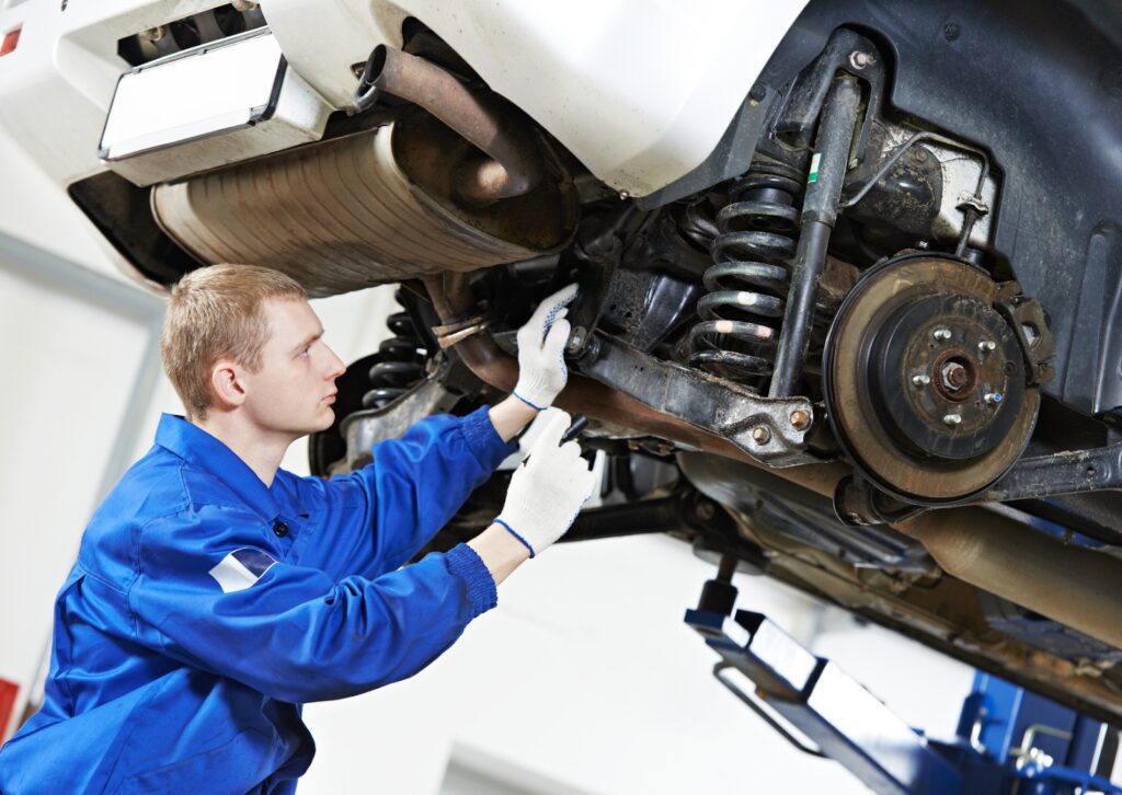 A mechanic in blue overalls inspects a car's steering and suspension system while working under a lifted vehicle.